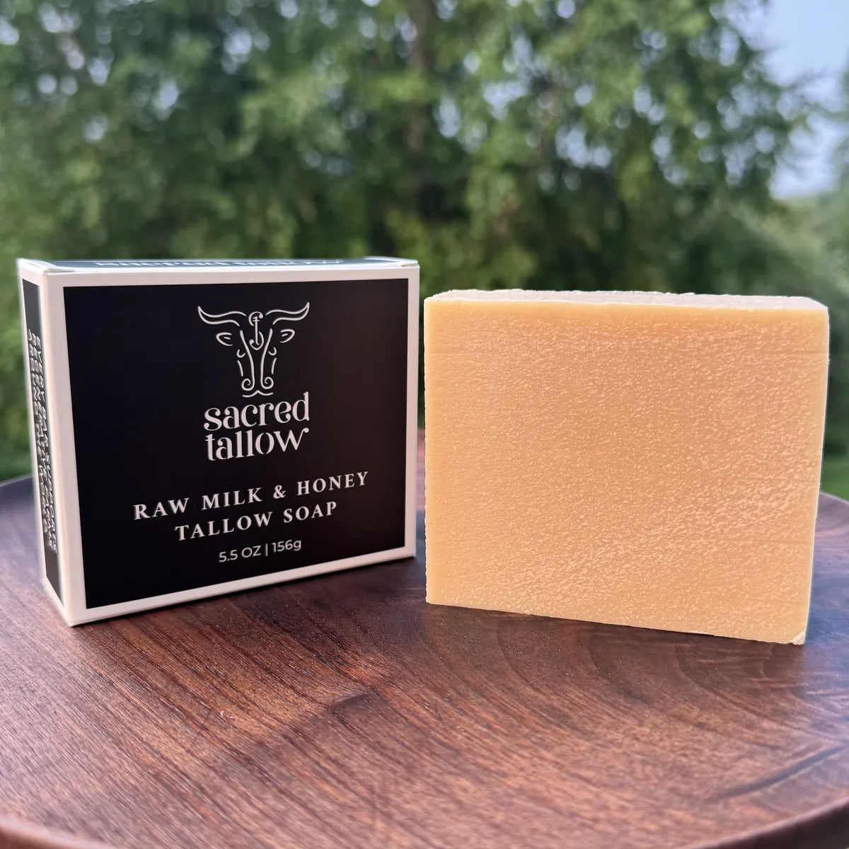 A bar of Sacred Tallow Raw Milk & Honey Tallow Soap stands next to its black and white packaging box on a round wooden table, with a blurred outdoor garden background.