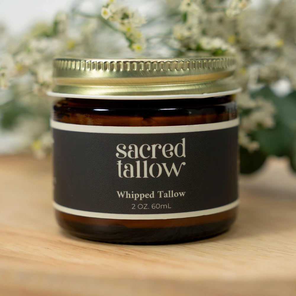 A jar of Sacred Tallow Whipped Tallow candle on a wooden surface with a blurred background of white flowers. The candle is in an amber glass jar with a gold metal lid and a black label with white text.