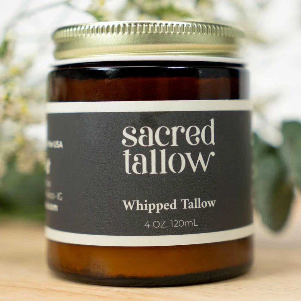 A 4 oz amber glass jar candle with a gold metal lid and a dark charcoal label reading 'sacred tallow Whipped Tallow 4 OZ. 120mL' on a light wooden surface with a soft, blurred background.