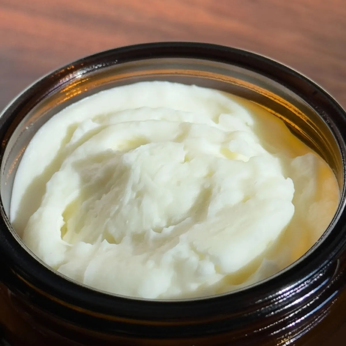 A close-up shot of a thick, creamy off-white cosmetic or skincare balm inside a dark amber glass jar, set against a warm brown wooden background.