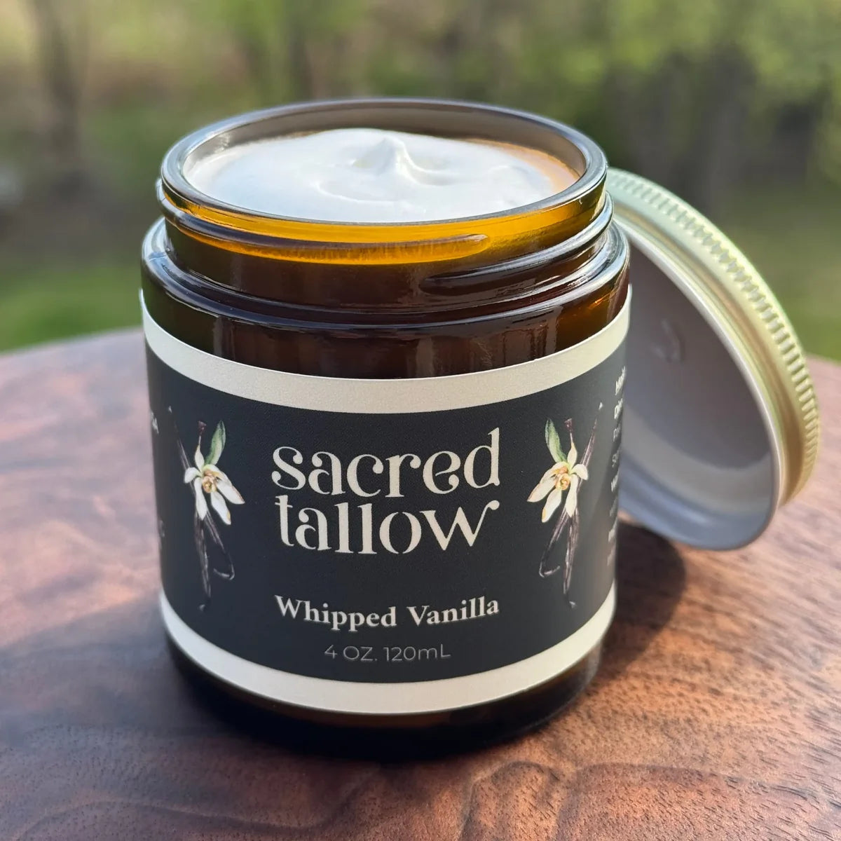 An open amber glass jar of Sacred Tallow whipped body butter on a wooden surface with a blurred outdoor background. The jar has a black label with white text and vanilla flower illustrations, and its gold lid rests beside it.