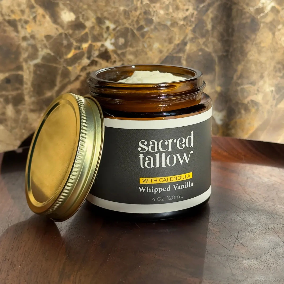 An open amber glass jar of Sacred Tallow body butter with a gold lid resting beside it on a dark wooden surface, set against a warm marble background. The label reads 'sacred tallow' and 'Whipped Vanilla'.