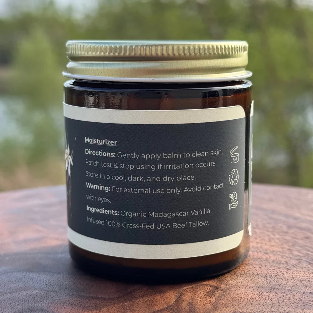 An amber glass jar of moisturizer with a gold metal screw lid and a black label, sitting on a wooden surface with a blurred outdoor background.